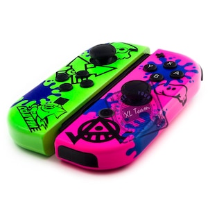 May include: A pair of custom Nintendo Switch Joy-Con controllers. The left Joy-Con is green with blue and black splatters and the text "SQUIDFORCE" on the side. The right Joy-Con is pink with black splatters and a black and white symbol.