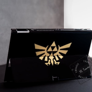 May include: Black Nintendo Switch console with a gold Triforce logo on the back. The console is in a stand and the XL Team logo is visible on the bottom right corner.