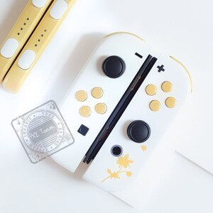 K&ouml;nnte beinhalten: Wei&szlig;e Nintendo Switch Joy-Con-Controller mit schwarzen Kn&ouml;pfen und goldenen Akzenten. Die Controller haben ein florales Design auf der rechten Seite.