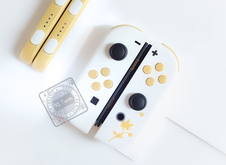 K&ouml;nnte beinhalten: Wei&szlig;e Nintendo Switch Joy-Con-Controller mit schwarzen Kn&ouml;pfen und goldenen Akzenten. Die Controller haben ein florales Design auf der rechten Seite.