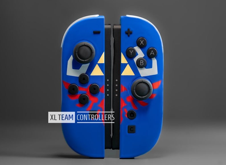 Puede incluir: Mandos Joy-Con XL TEAM CONTROLLERS azules y blancos con detalles en rojo y dorado. Los mandos tienen botones y joysticks negros, con el texto "XL TEAM CONTROLLERS" en blanco.