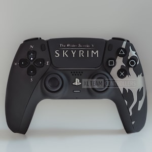 May include: A black PlayStation 5 controller with a custom design featuring a white dragon and the text "The Elder Scrolls V Skyrim" and "XL Team Controllers".