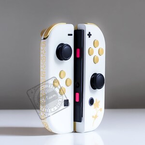K&ouml;nnte beinhalten: Ein wei&szlig;er Nintendo Switch Joy-Con-Controller mit goldenen Akzenten und schwarzen Kn&ouml;pfen. Der Controller hat ein goldenes Muster an der Seite und ein goldenes Ahornblatt-Design auf der R&uuml;ckseite.