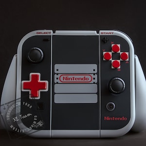 May include: A black and white Nintendo Switch console with a custom NES design. The console has red buttons and a red plus button. The console has the text "Nintendo" on the front.