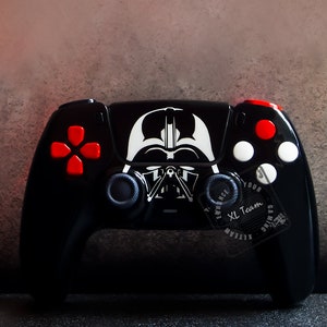 May include: A black PlayStation 5 controller with a Darth Vader design. The controller has red and white buttons and a black and white sticker of Darth Vader's face on the front. The text "Enhance Your Gaming XL Team" is printed on the back of the controller.