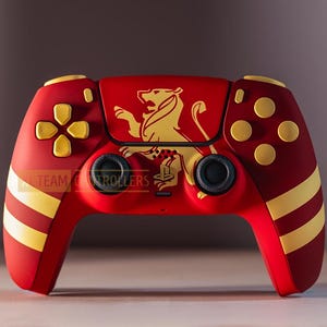 May include: A red and gold PlayStation 5 controller with a lion crest design. The controller has a red base with gold stripes and a gold lion crest on the front. The controller has black buttons and triggers.