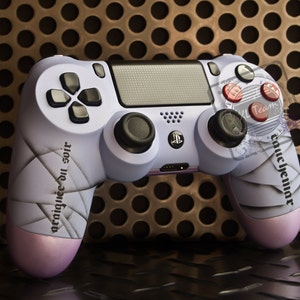 May include: A purple and white PlayStation 4 controller with a custom design featuring a white, geometric pattern and the text "araignée du soir" on the left side. The controller has red buttons and a black touchpad.