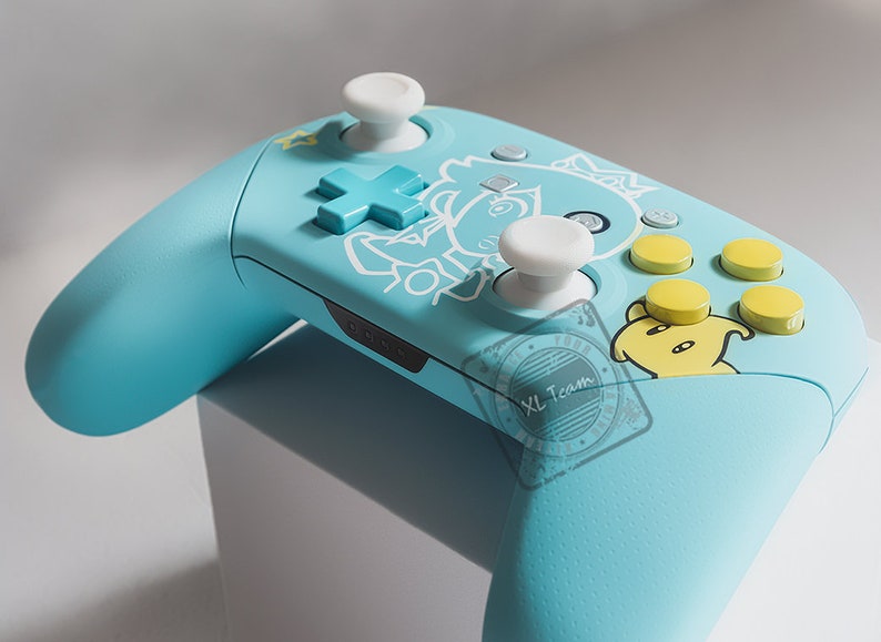 May include: A light blue video game controller with a white and yellow design. The controller has a white joystick, a white directional pad, and four yellow buttons. The controller is decorated with a cartoon character.