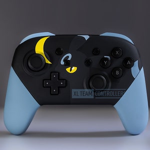 May include: A black Nintendo Switch controller with a blue and yellow design featuring a cartoon cat with a crescent moon. The text "XL TEAM CONTROLLERS" is printed on the controller.