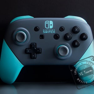 May include: A teal and gray Nintendo Switch controller with the text "NINTENDO SWITCH" on the front. The controller has a circular button with a white border on the right side and a similar button on the left side. The controller has a black D-pad and four face buttons labeled "A", "B", "X", and "Y". The controller has a teal and gray stamp on the right side with the text "XL Team" in a circle.