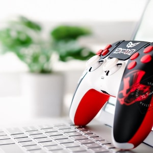 May include: A white, black, and red gaming controller with the text "Play Your Part" and a red graphic design. The controller is resting on a laptop keyboard, with a blurred background of a plant in a white pot.