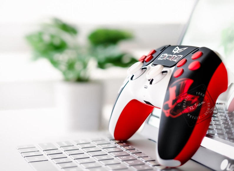 May include: A white, black, and red gaming controller with the text "Play Your Part" and a red graphic design. The controller is resting on a laptop keyboard, with a blurred background of a plant in a white pot.