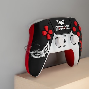 May include: A custom video game controller with a black, red, and white color scheme. The controller features a design with the text "TAKE YOUR HEART" and a stylized mask. The buttons are red, and the joysticks are white.