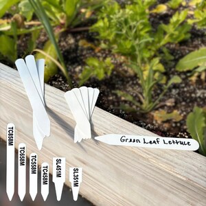 May include: A collection of white, pointed plant markers, some blank and some featuring black printed text including TC858M, TC658M, TC558M, TC458M, SL451M, SL351M, and one horizontal marker labeled Green Leaf Lettuce, arranged on a wooden surface.