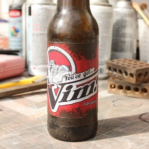 以下が含まれることがあります： 赤、白、黒のラベルが付いた茶色のガラス瓶。ラベルには、赤いジャンプスーツを着た漫画の女性と「You've got... Vim!」という文字が書かれている。瓶は市松模様の表面の上に置かれている。