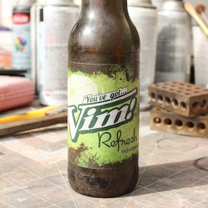 Puede incluir: Una botella de vidrio marrón con una etiqueta verde y blanca que dice "You've got... Vim! Refresh". La botella está sobre una superficie a cuadros.