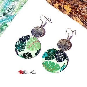 Puede incluir: Un par de pendientes colgantes con un diseño de hojas tropicales. Los pendientes están hechos de metal y tienen un acabado plateado. La parte superior de los pendientes es un disco redondo plateado con un diseño decorativo. La parte inferior de los pendientes es un disco redondo verde con un diseño de hojas tropicales.