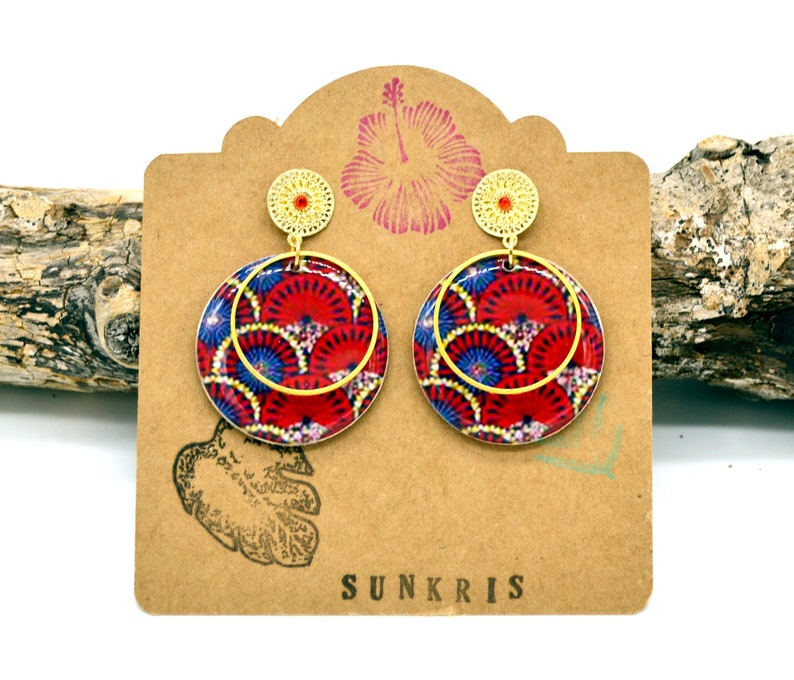 May include: Gold dangle earrings with a red, blue, and white patterned circle design. The earrings are displayed on a brown card with the word "SUNKRIS" printed on it.