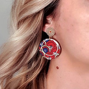 May include: A pair of round earrings with a red, blue, and white patterned design. The earrings have a gold-colored metal frame and a small gold-colored circle at the top.