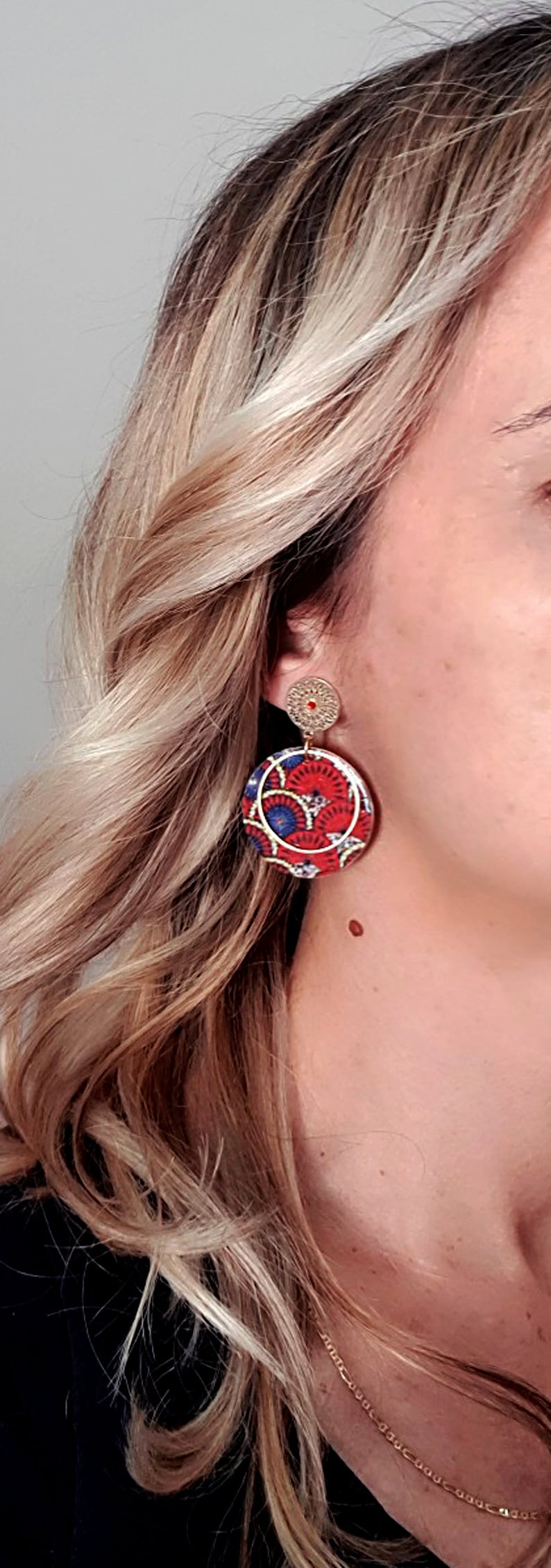 May include: A pair of round earrings with a red, blue, and white patterned design. The earrings have a gold-colored metal frame and a small gold-colored circle at the top.