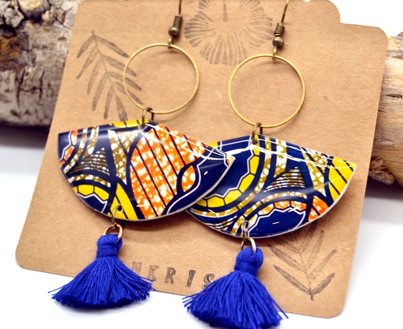 May include: A pair of blue and yellow patterned earrings with gold hoops and blue tassel accents. The earrings are on a brown card with the word "NKRIS" printed on it.
