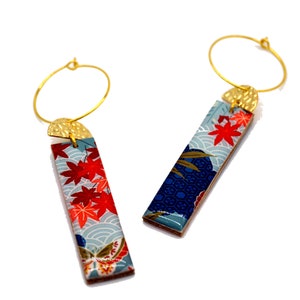 May include: A pair of gold hoop earrings with rectangular pendants. The pendants feature a Japanese-inspired design with red maple leaves, blue waves, and gold accents. The earrings are set against a white background.