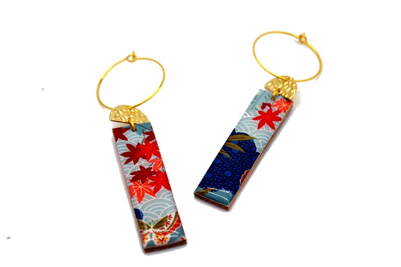 May include: A pair of gold hoop earrings with rectangular pendants. The pendants feature a Japanese-inspired design with red maple leaves, blue waves, and gold accents. The earrings are set against a white background.