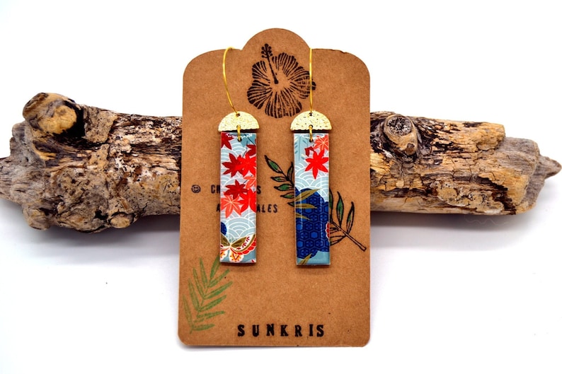 May include: A pair of rectangular earrings with a Japanese-inspired design. The earrings feature a gold-colored top and a colorful pattern of red maple leaves and blue waves. The earrings are displayed on a brown card with the word "SUNKRIS".