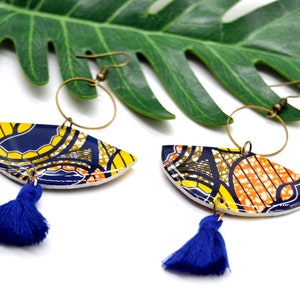 May include: A pair of colorful earrings with a blue tassel. The earrings are made of a white base with a yellow, orange, and blue patterned design. The earrings are hanging from gold hoops.