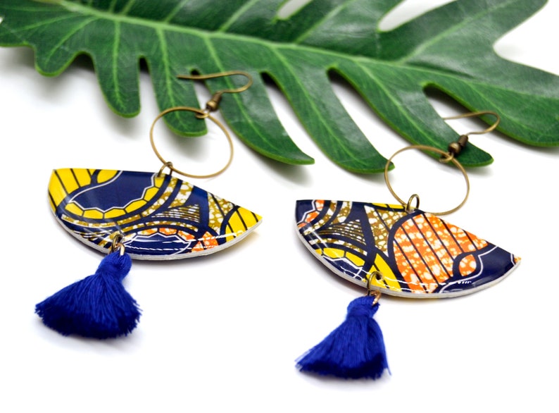 May include: A pair of colorful earrings with a blue tassel. The earrings are made of a white base with a yellow, orange, and blue patterned design. The earrings are hanging from gold hoops.