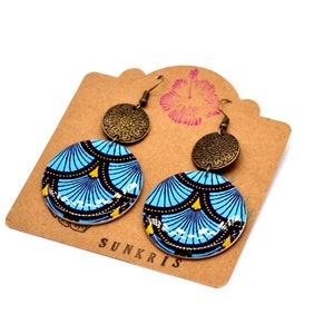 May include: A pair of blue and gold patterned earrings with a bronze charm. The earrings are on a brown card with the word "SUNKRIS" printed on it.