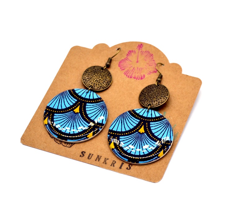 May include: A pair of blue and gold patterned earrings with a bronze charm. The earrings are on a brown card with the word "SUNKRIS" printed on it.