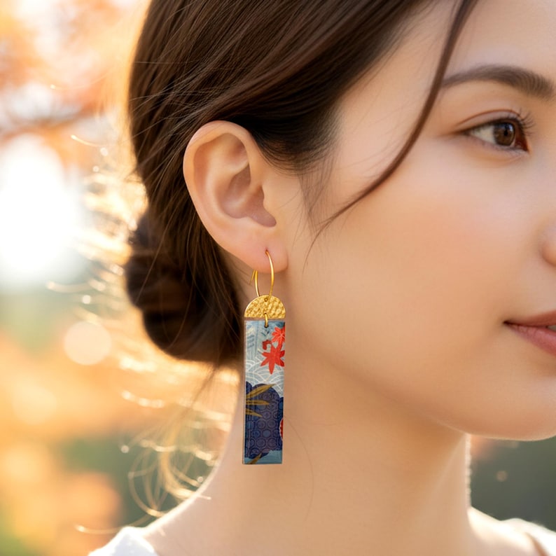 Gros plan d'une boucle d'oreille rectangulaire en papier japonais bleu foncé et rouge, motifs vagues et érable, suspendue à une attache dorée martelée.