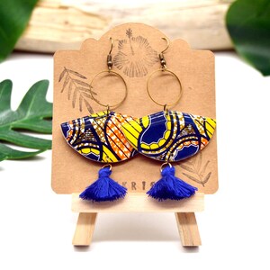May include: A pair of blue and yellow patterned earrings with gold hoops and blue tassels. The earrings are displayed on a wooden easel.