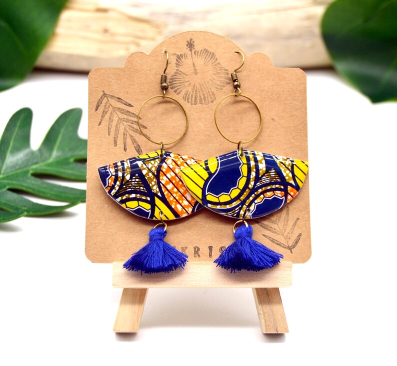 May include: A pair of blue and yellow patterned earrings with gold hoops and blue tassels. The earrings are displayed on a wooden easel.