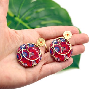 May include: A pair of red and blue patterned circle earrings with gold accents. The earrings have a gold hoop and a smaller gold circle with a red jewel in the center.
