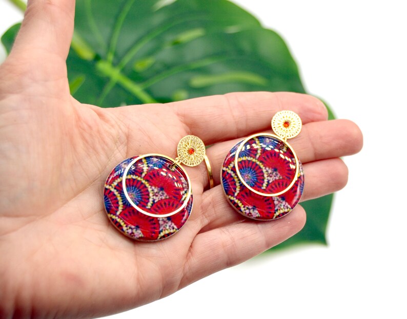 May include: A pair of red and blue patterned circle earrings with gold accents. The earrings have a gold hoop and a smaller gold circle with a red jewel in the center.