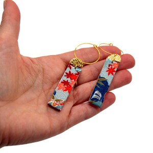 May include: A pair of gold hoop earrings with rectangular pendants. The pendants feature a light blue background with red maple leaf and wave patterns. The top of each pendant has a gold-colored cap.