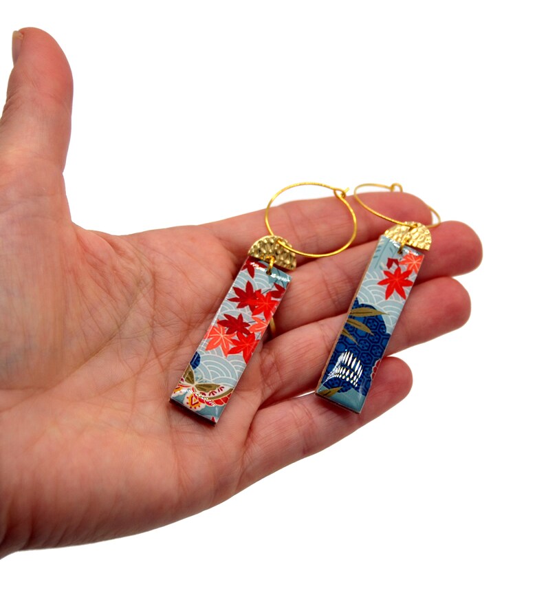 May include: A pair of gold hoop earrings with rectangular pendants. The pendants feature a light blue background with red maple leaf and wave patterns. The top of each pendant has a gold-colored cap.