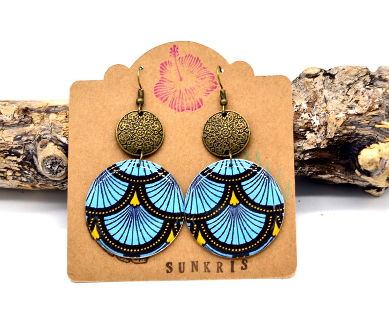 May include: A pair of blue and gold patterned earrings with a bronze filigree charm. The earrings are hanging on a brown card with the word "SUNKRIS" printed on it.