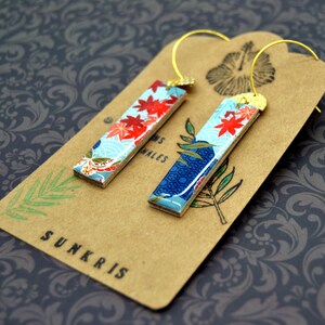May include: A pair of rectangular earrings with a Japanese-inspired design. The earrings feature a light blue background with red floral accents and gold-colored ear wires. They rest on a brown tag with the word "SUNKRIS".