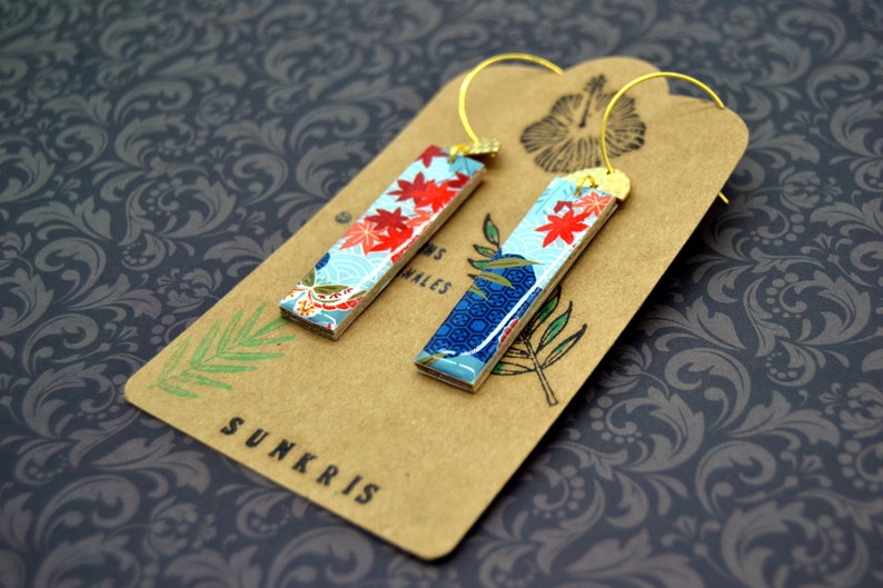 May include: A pair of rectangular earrings with a Japanese-inspired design. The earrings feature a light blue background with red floral accents and gold-colored ear wires. They rest on a brown tag with the word "SUNKRIS".