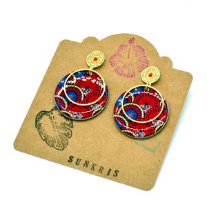 May include: A pair of gold hoop earrings with red and blue patterned circles. The earrings are on a brown card with the word "SUNKRIS" printed on it.