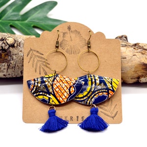 May include: A pair of blue and yellow patterned earrings with gold hoops and blue tassels. The earrings are displayed on a brown card with the word "KRIS" printed on it.