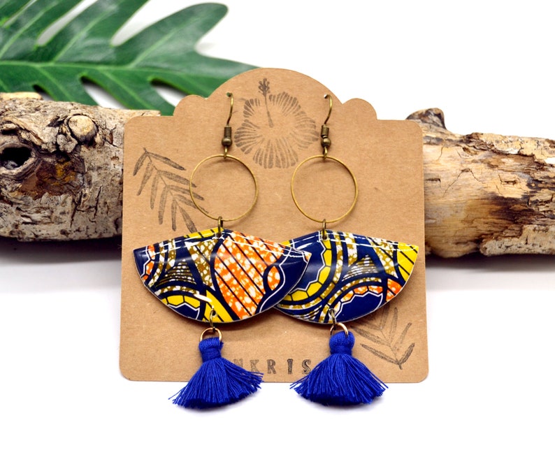 May include: A pair of blue and yellow patterned earrings with gold hoops and blue tassels. The earrings are displayed on a brown card with the word "KRIS" printed on it.