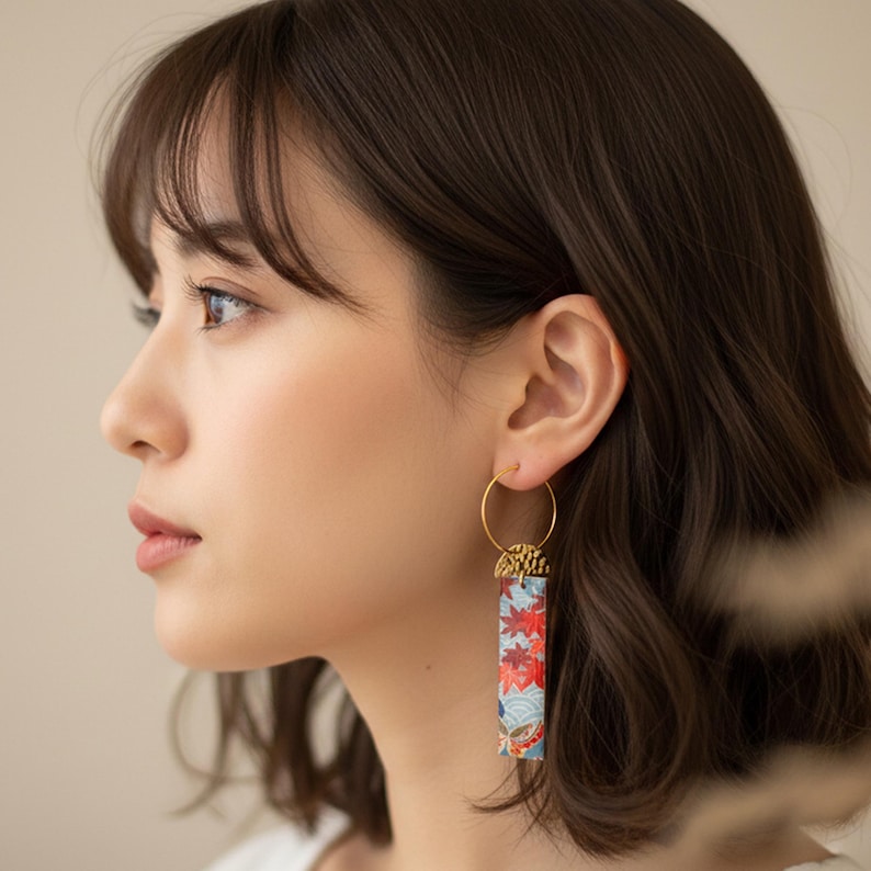 May include: Gold hoop earrings with a rectangular, patterned drop. The drop features a floral design with red, orange, and blue hues. The earrings are worn by a person with dark hair.