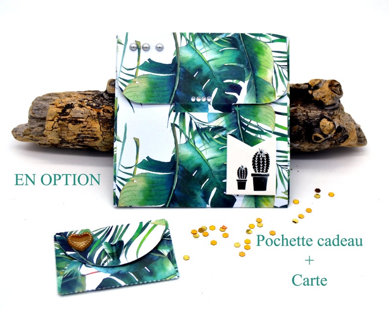 May include: A white gift bag with a tropical leaf pattern and a black cactus graphic. The bag is closed with a flap and has silver metal accents. The text "EN OPTION" is visible in the image. The bag is sitting on a piece of driftwood.
