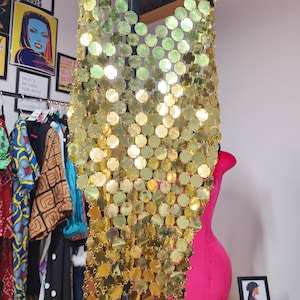 May include: A sleeveless top constructed from numerous small, reflective gold-colored circular metal discs linked together in a chainmail pattern. The garment features a halter-style neckline and a semi-sheer, draped design that hangs vertically.