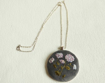 Puede incluir: Un collar de cadena plateada con un colgante redondo gris con flores bordadas en rosa y blanco.