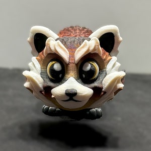 Peut inclure: Une figurine de panda roux imprimée en 3D avec de grands yeux, un museau blanc et une fourrure brune. La figurine est peinte avec un dégradé de brun, rouge et blanc.